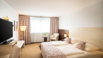 A hotel room with a bed, a desk with a TV, a chair, a table with a plant, and curtains.