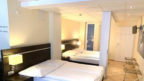 Habitación de hotel con dos camas, una mesita de noche, una lámpara, un televisor montado en la pared y una puerta que da a otra habitación.