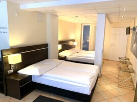 Habitación de hotel con dos camas, una mesita de noche, una lámpara, un televisor montado en la pared y una puerta que da a otra habitación.