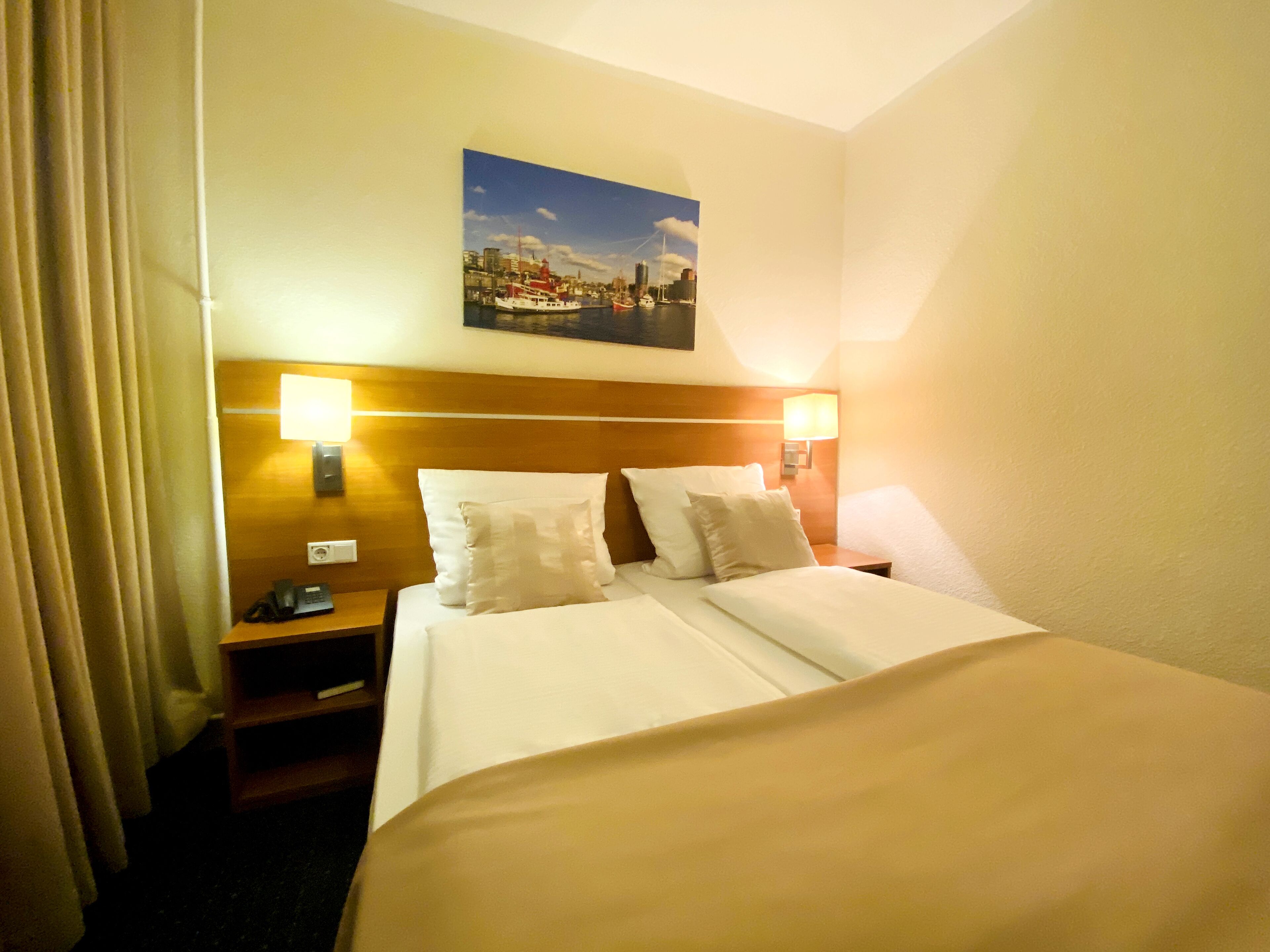 A hotel room with a large bed, two bedside tables, a nightstand, a painting of a cityscape, and a curtain.