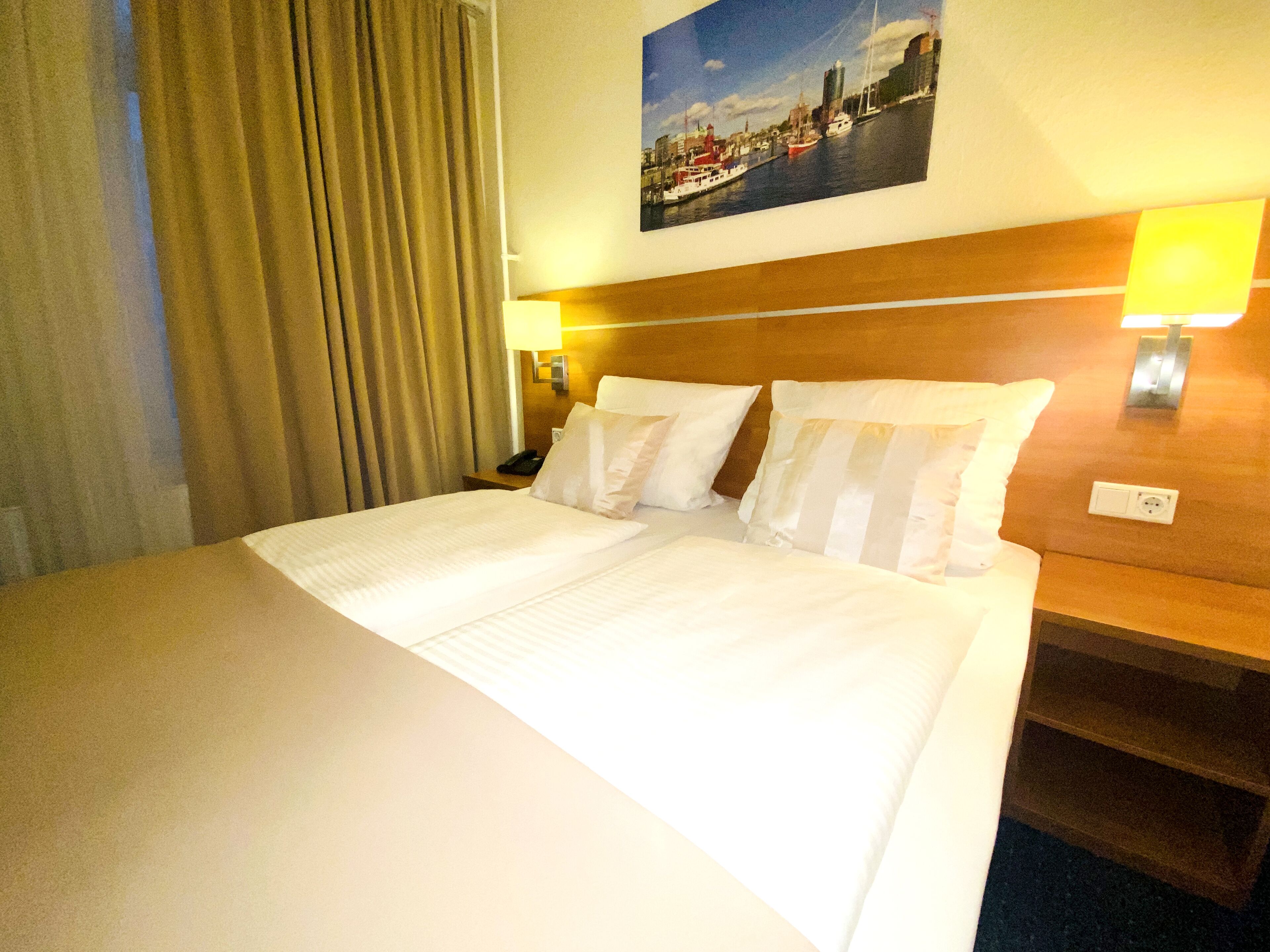A hotel room with a large bed, two bedside lamps, a headboard, a nightstand, and a painting of a cityscape.