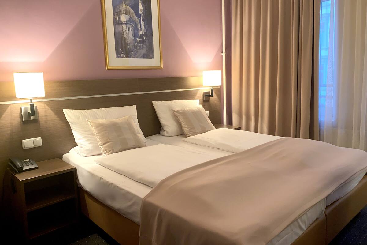A hotel room with a bed, two bedside lamps, a nightstand with a phone, and a painting on the wall.
