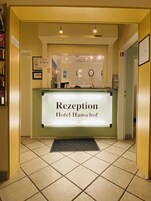 Ein Empfangstresen mit einem Schild, auf dem 'Rezeption Hotel Hansehof' steht, in der Hotelhalle.