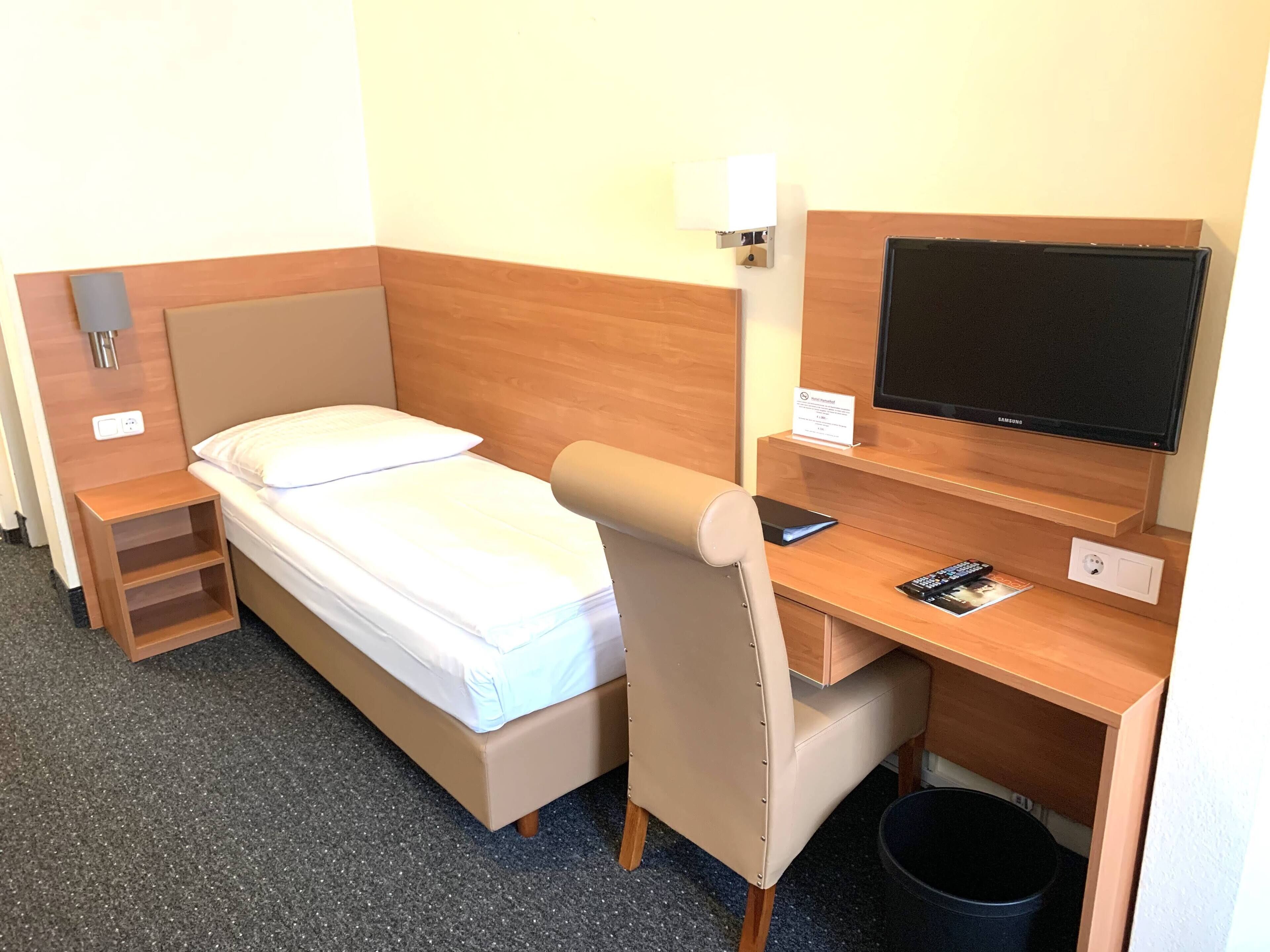 A hotel room with a bed, a desk with a computer, a TV, and a trash bin.