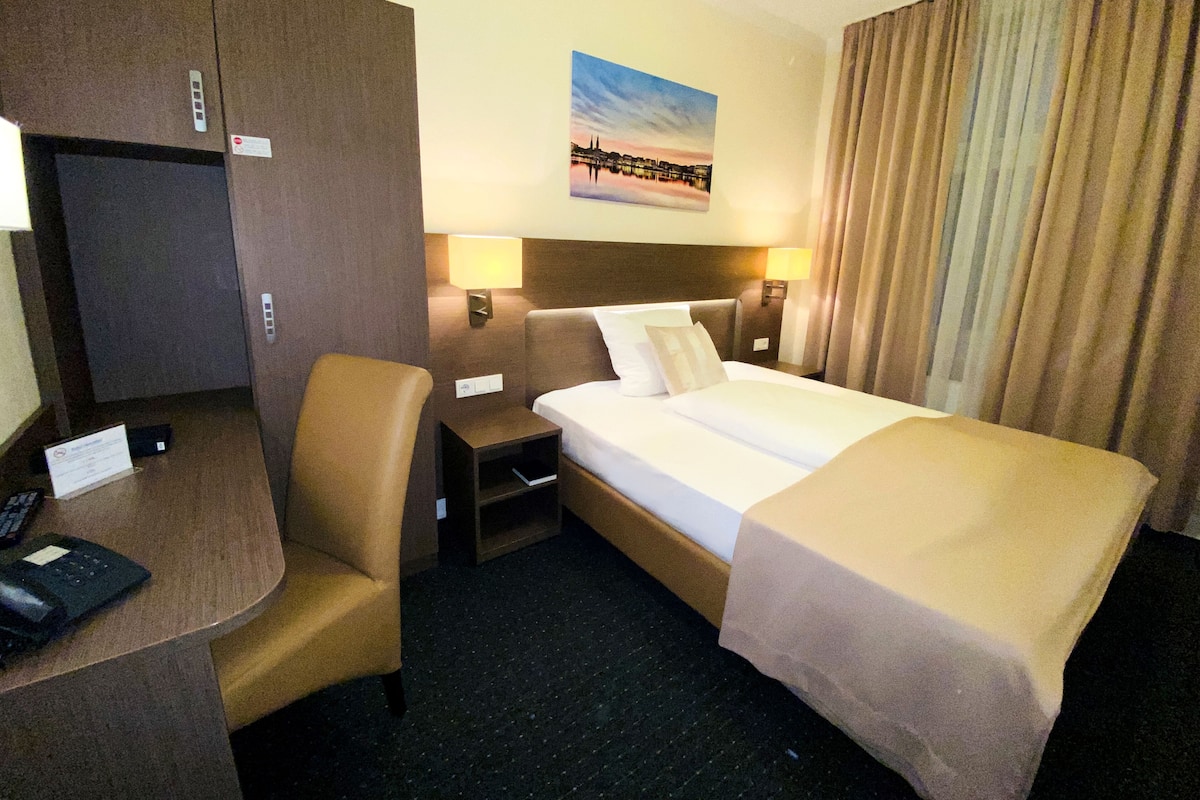 A hotel room with a bed, a desk, a chair, a television, and a painting on the wall.