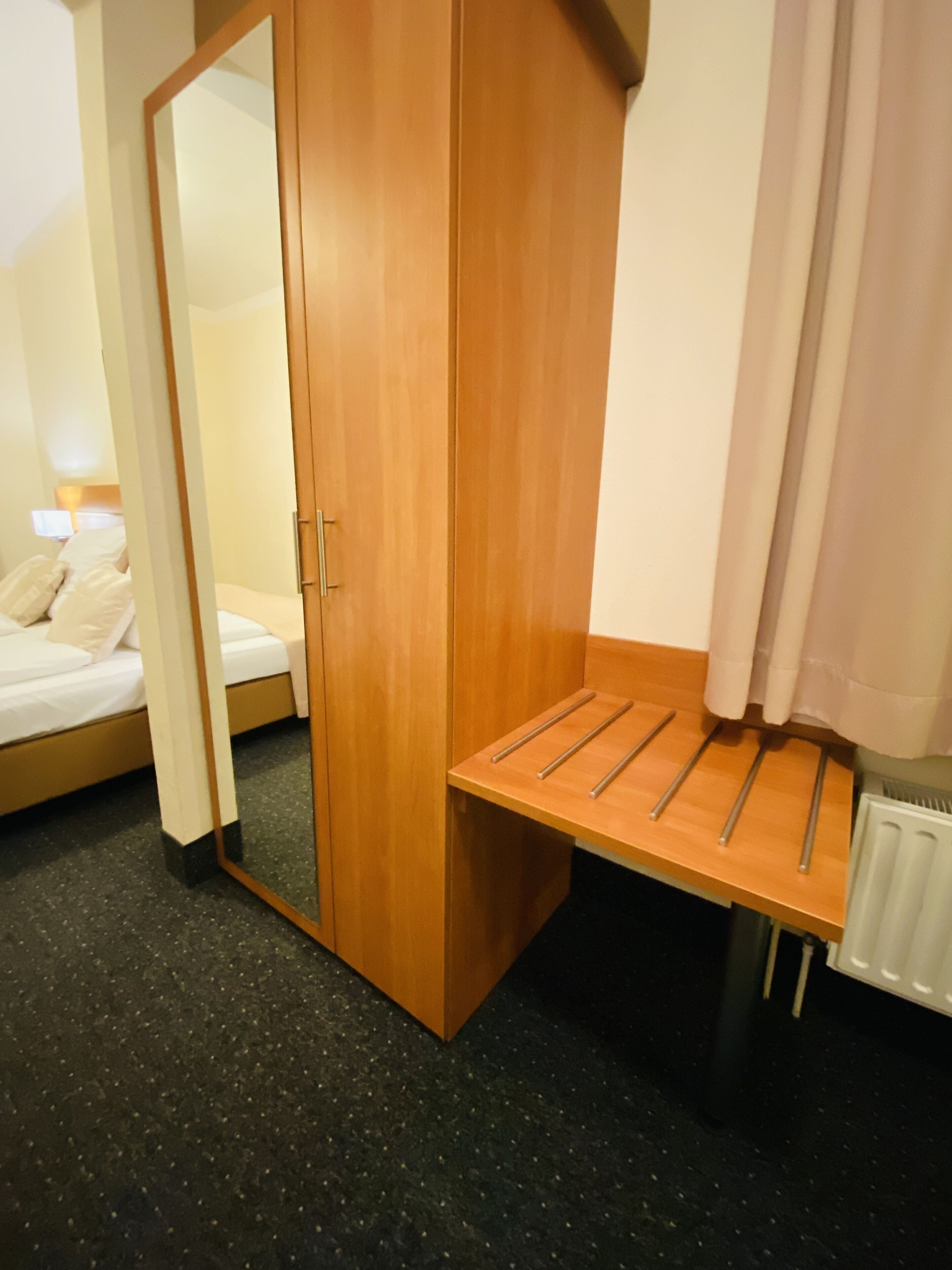 Ein Hotelzimmer mit einem Holzschrank, einer kleinen Holzbank mit Metallstreben, einem Bett mit weißer Bettwäsche und einem Heizkörper am Boden.