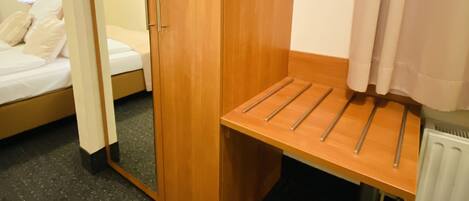 Ein Hotelzimmer mit einem Holzschrank, einer kleinen Holzbank mit Metallstreben, einem Bett mit weißer Bettwäsche und einem Heizkörper am Boden.