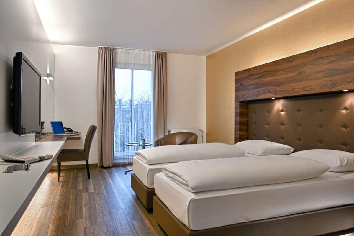 A hotel room with two beds, a desk with a laptop, a chair, a TV, and a large window with curtains.