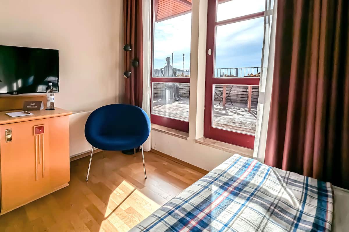 Quarto de hotel com uma cama, uma escrivaninha com televisão, uma cadeira e uma janela com vista para um cais.