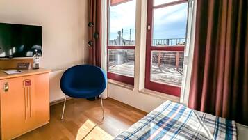 A hotel room with a bed, a desk with a television, a chair, and a window with a view of a pier.