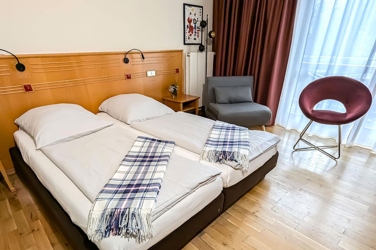 Uma cama de casal com manta xadrez, uma poltrona cinza e uma cadeira vermelha em um quarto com piso de madeira e uma janela com cortinas.