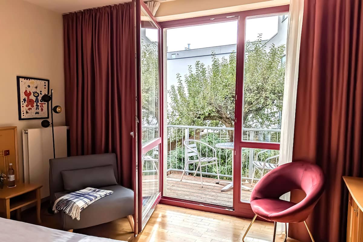 Quarto com sofá, uma cadeira vermelha, uma mesa e varanda com guarda-corpo.