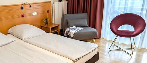 Habitación de hotel con dos camas, una pequeña mesa con un florero, un sillón gris, una silla roja y una ventana con cortinas.
