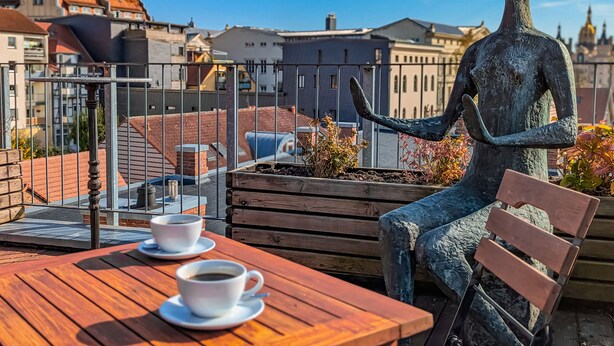 Een standbeeld van een zittende figuur met een kopje en schoteltje op een houten tafel, met uitzicht op een stadsgezicht met gebouwen en een heldere lucht.