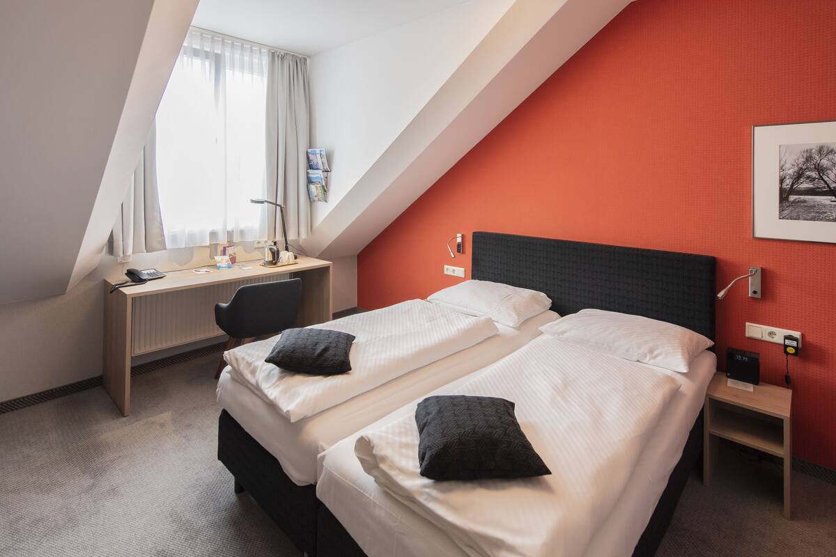 Quarto de hotel com duas camas, uma mesa e janela com cortinas.