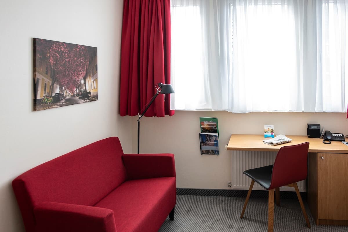 Quarto com sofá vermelho, escrivaninha de madeira com cadeira, radiador e janela com cortinas vermelhas.