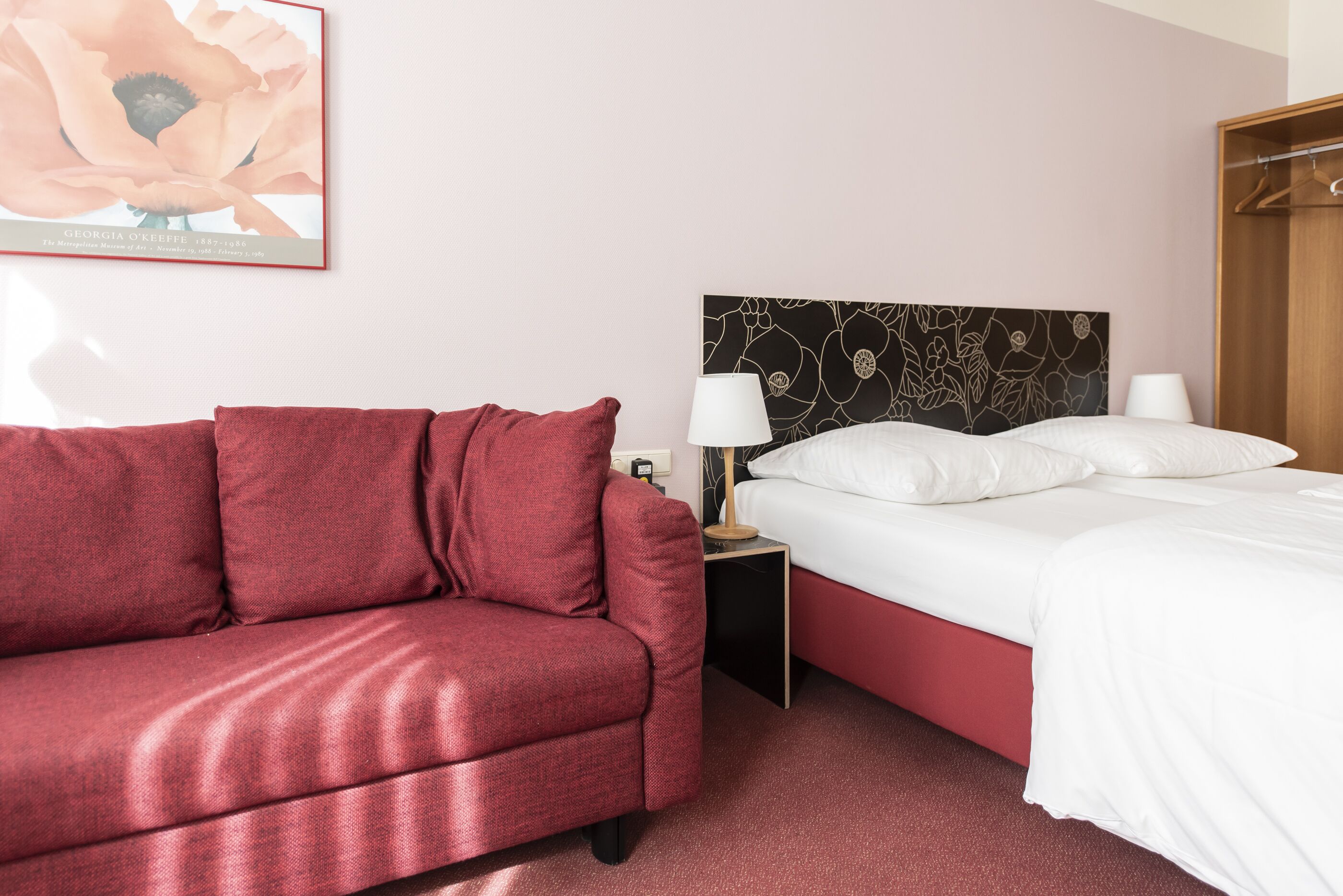 A hotel room with a red sofa, a bed with white bedding, a nightstand with a lamp, and a framed artwork on the wall.