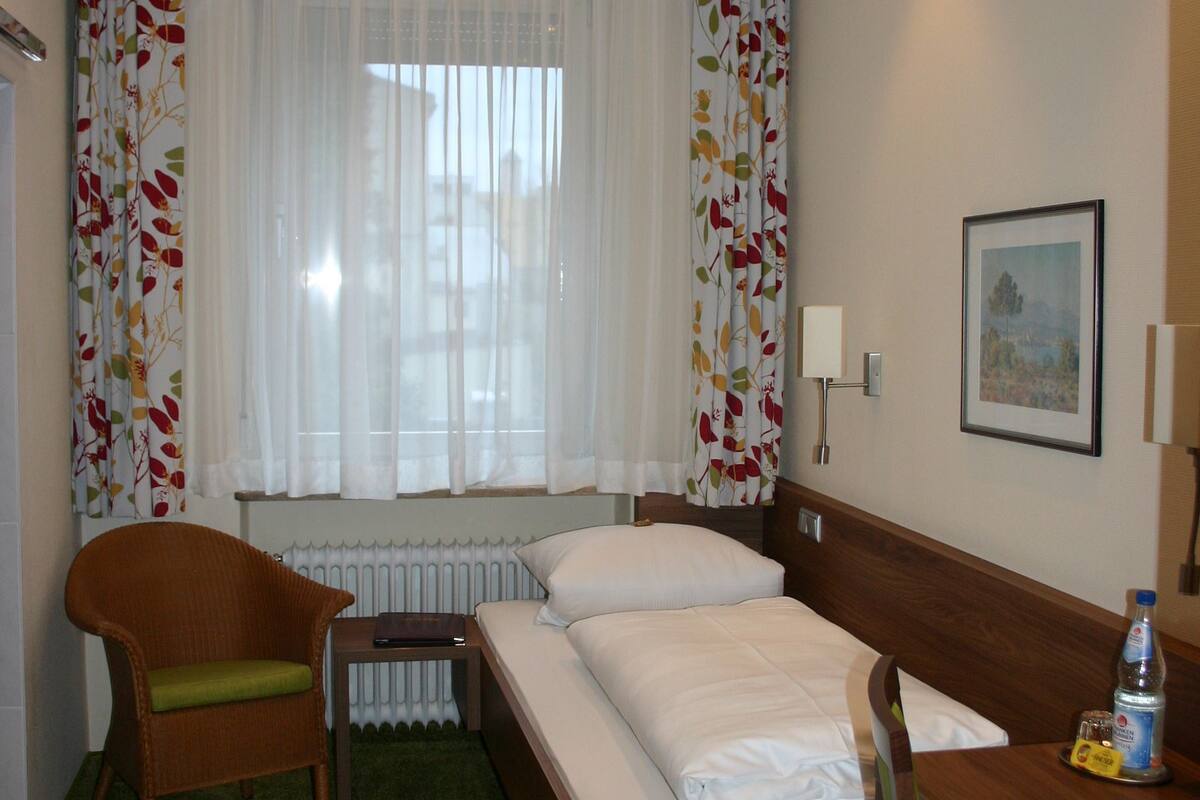 A hotel room with a bed, a desk, a chair, a radiator, and a window with curtains.