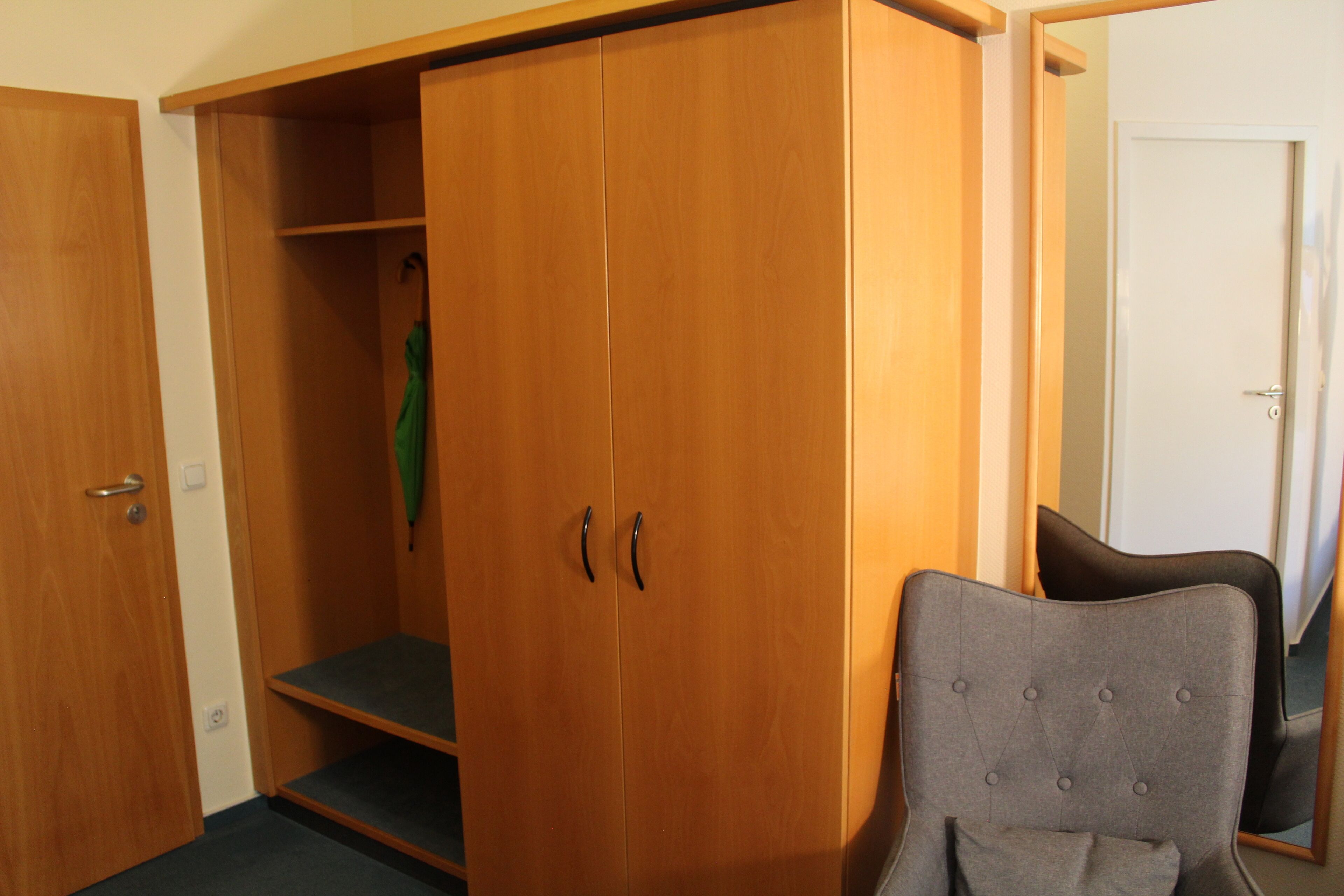 Een kamer met een houten garderobe, een grijs gestoffeerde fauteuil en een spiegel die een deel van de kamer weerspiegelt.