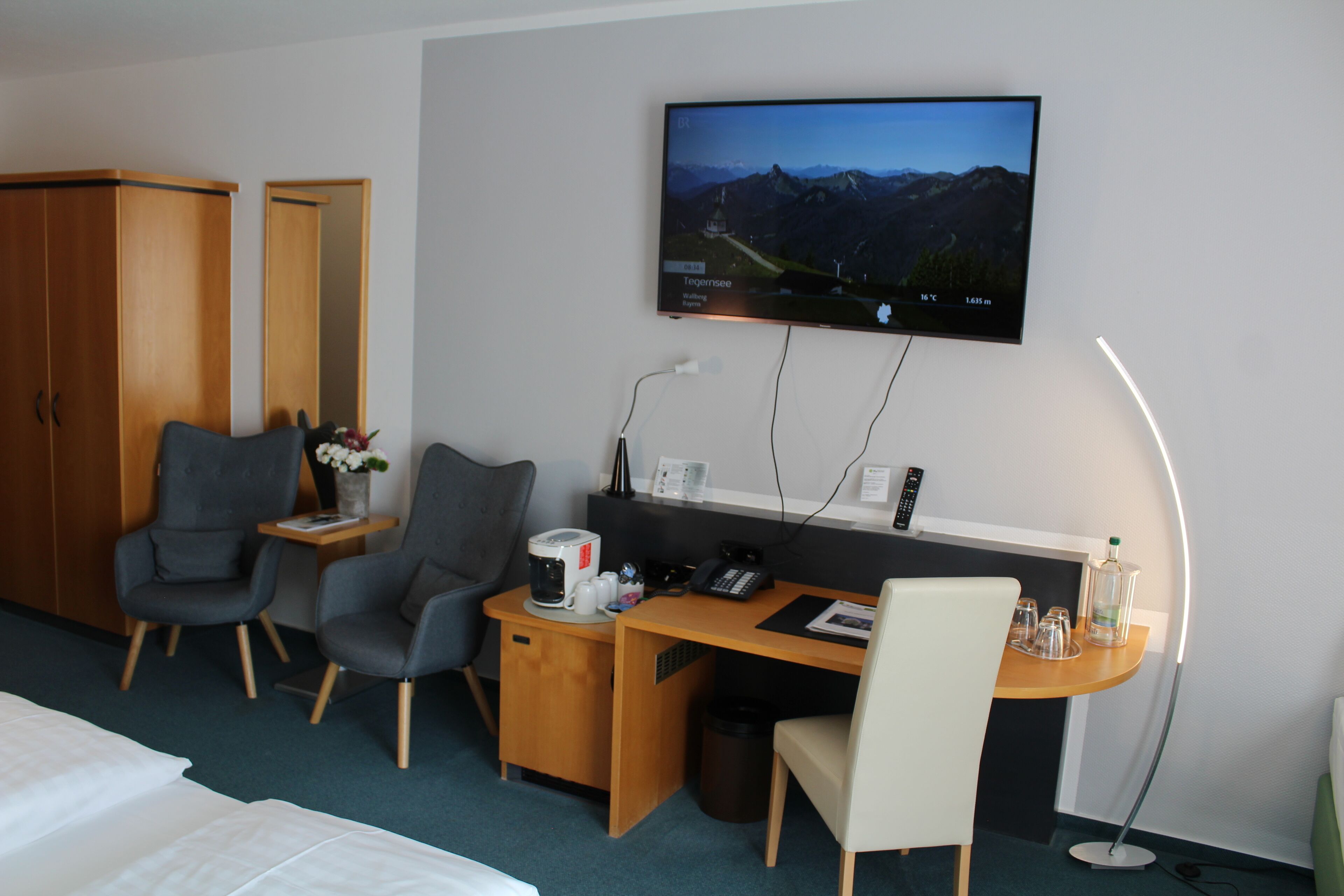 Habitación de hotel con cama, dos sillones, escritorio con computadora, televisor montado en la pared y una mesita de noche con lámpara.