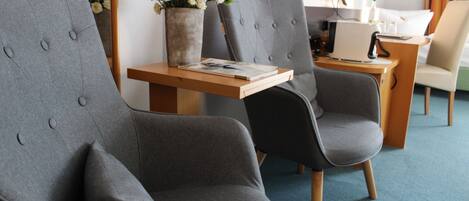 Et moderne hotelværelse med grå polstrede stole, et træsidebord med en bog og en vase med blomster, samt et skrivebord med en computer og en lampe.