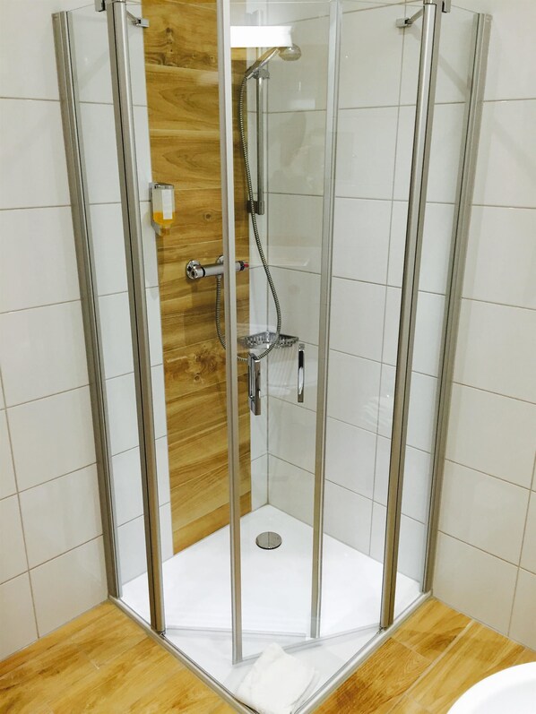 A glass-enclosed shower stall with a wooden panel wall, a white tiled floor, and a metal showerhead.
