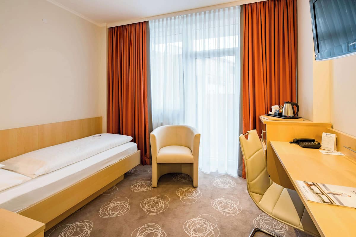 Habitació d'hotel amb un llit individual, una cadira, un escriptori i una finestra amb cortines.