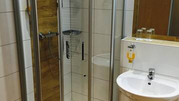 A glass-enclosed shower with a wooden panel wall, a white sink with a mirror above it, and a small trash bin next to the sink.