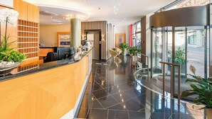A hotel reception area with a modern design, including a reception desk, seating area, and a glass elevator.