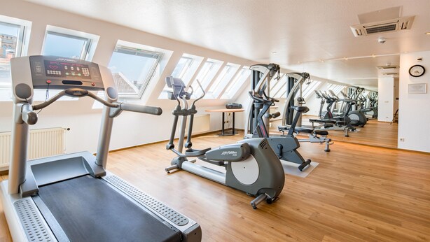 Un gimnasio con varias máquinas de ejercicio, amplias ventanas y suelo de madera.