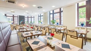 A restaurant with multiple tables and chairs, a long bench seating area, and large windows providing natural light.