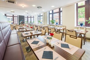 Un restaurante con varias mesas y sillas, una zona de estar con bancos largos, y amplias ventanas que aportan luz natural.