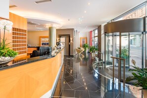 Un lobby moderno de hotel con una recepción, zona de estar y un ascensor de vidrio.