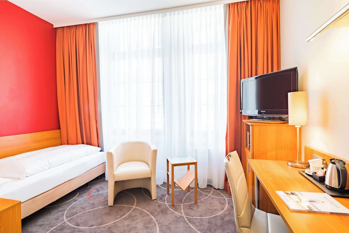 Habitació d'hotel amb un llit, una cadira, una taula petita, una televisió i un escriptori amb una làmpada i una cafetera.
