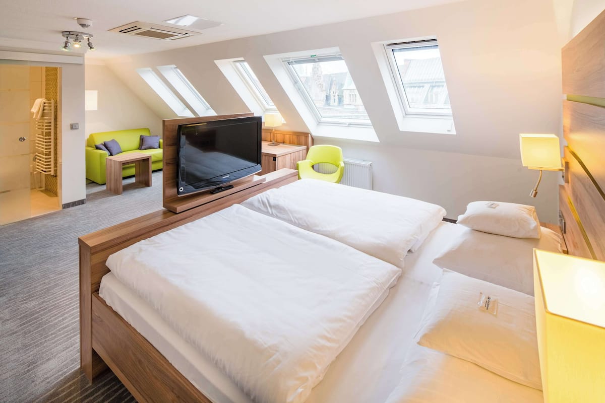 Habitació d'hotel amb dos llits, televisió, un sofà verd i finestres velàries.