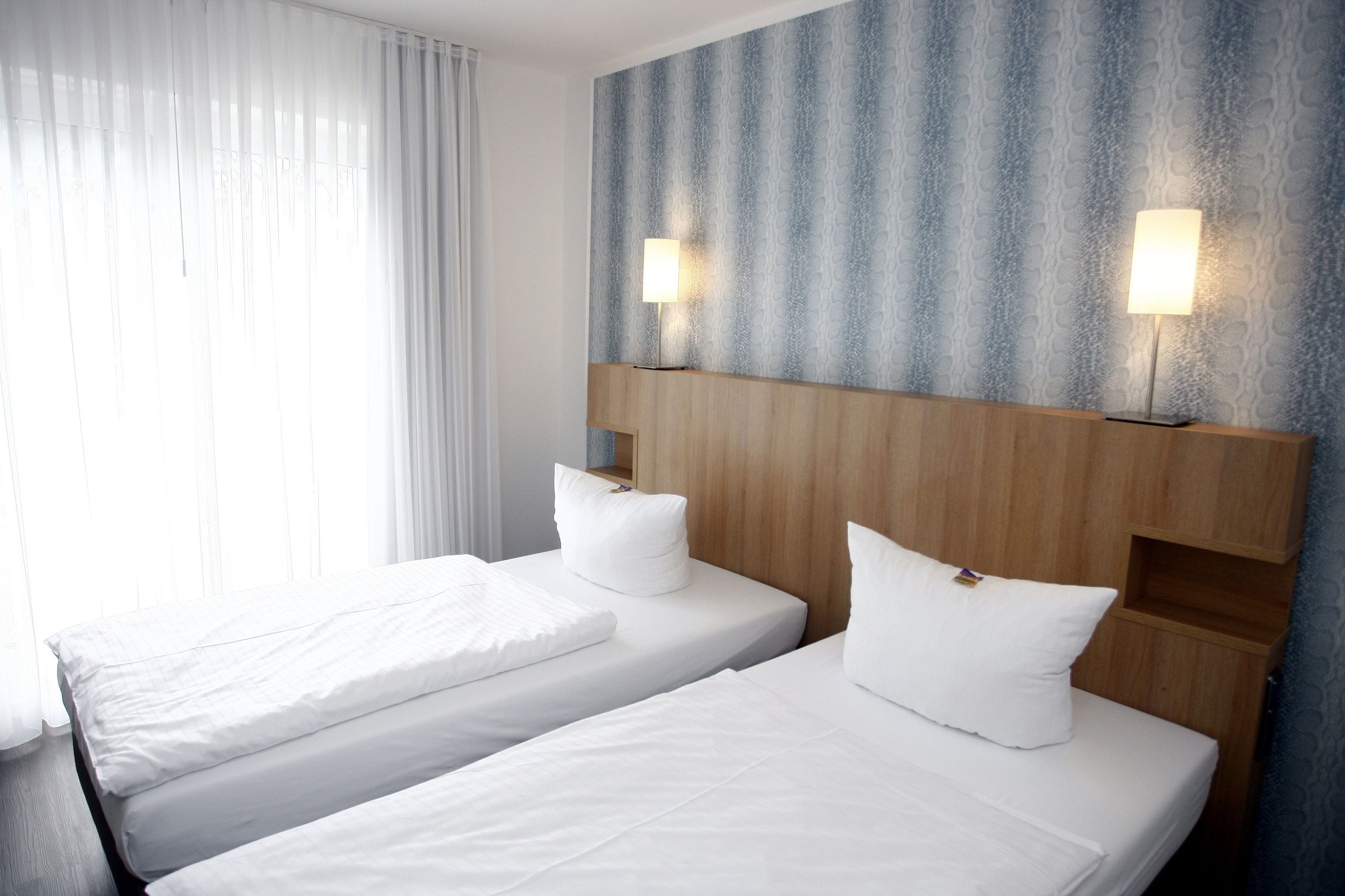 Habitación de hotel con dos camas, ropa de cama blanca, cabecero de madera y pared con diseño.