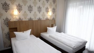 Une chambre d’hôtel avec deux lits, une tête de lit en bois, un papier peint à motifs et une fenêtre avec des rideaux légers.