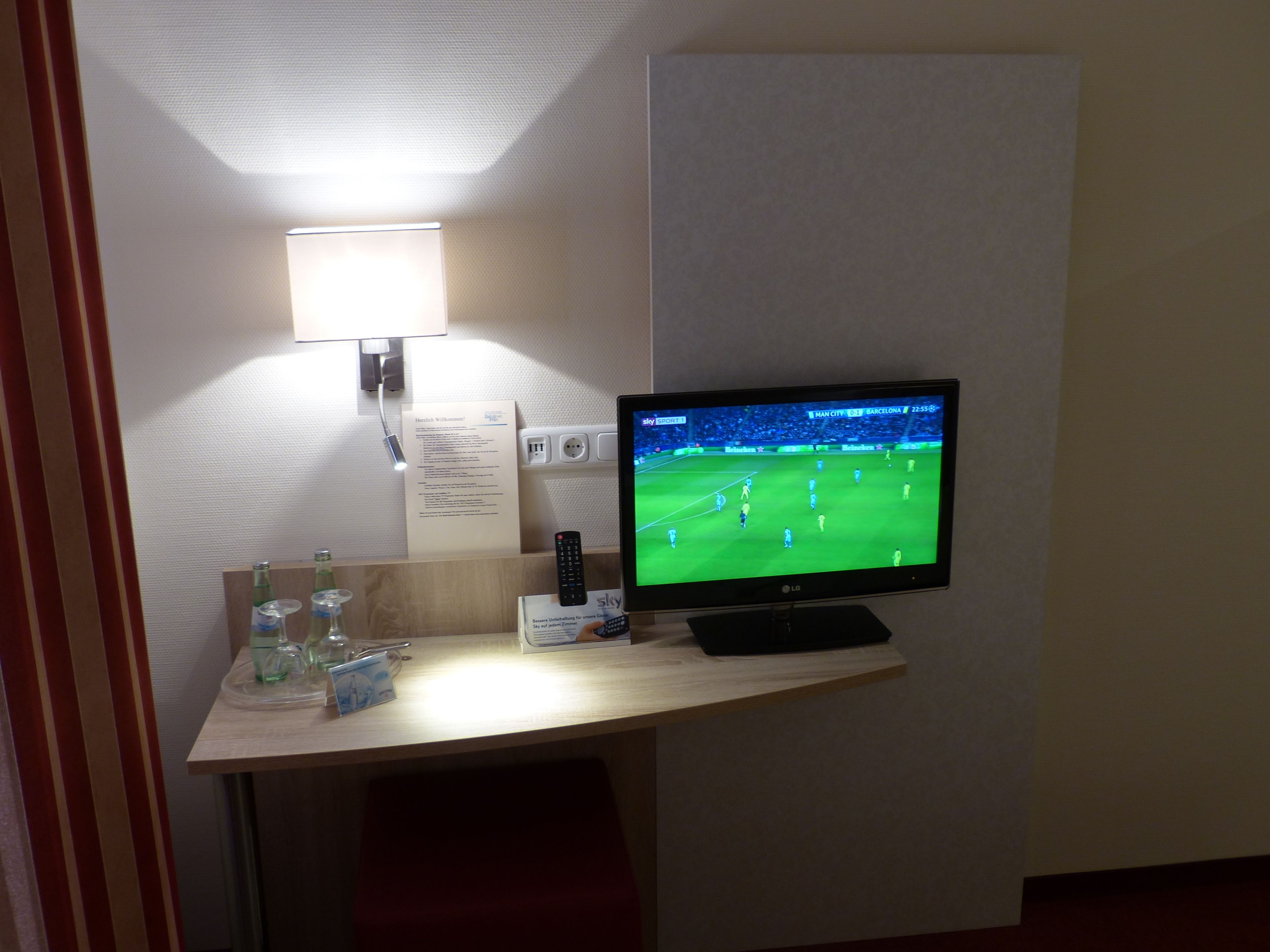 A hotel room with a flat-screen TV mounted on a wall, a bedside table with a lamp, and a small shelf with bottles and glasses.