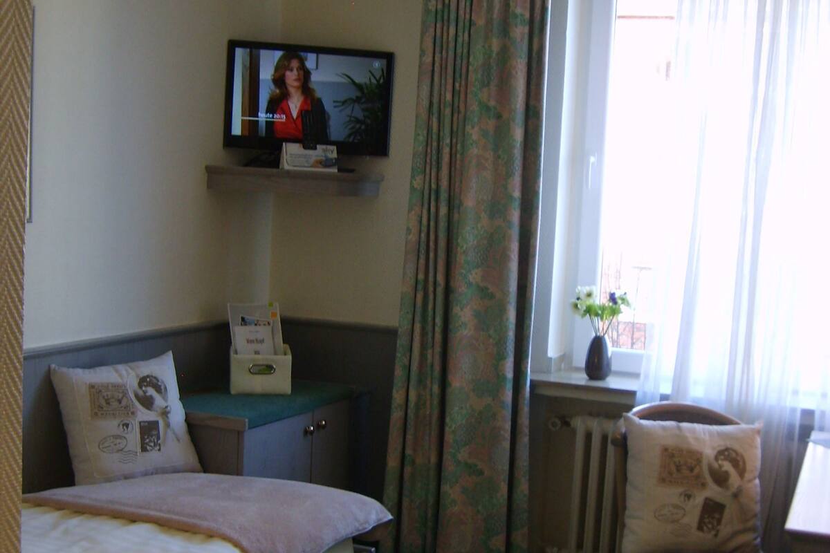 A hotel room with a bed, a television mounted on the wall, a desk with a chair, and a window with curtains.