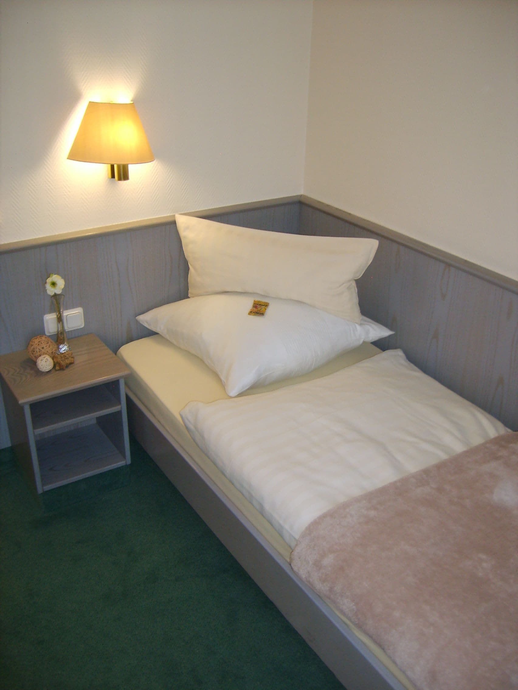 A single bed with a wooden nightstand, a lamp, and a small vase with a flower.
