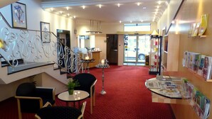 A hotel lobby with a red carpet, a reception desk, a staircase with metal railings, and a wall with magazines. - City Hotel by Celina (Hagen)