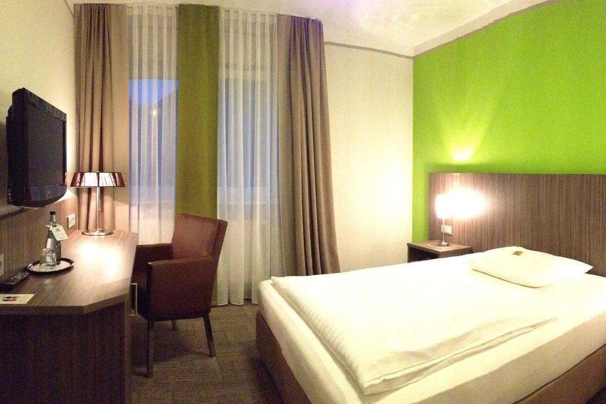 A hotel room with a bed, a desk with a lamp, a chair, a TV, and a nightstand with a phone.