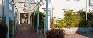 A covered walkway with a glass roof, flanked by plants and a concrete wall. - Hotel Ambiente Walldorf (Walldorf)