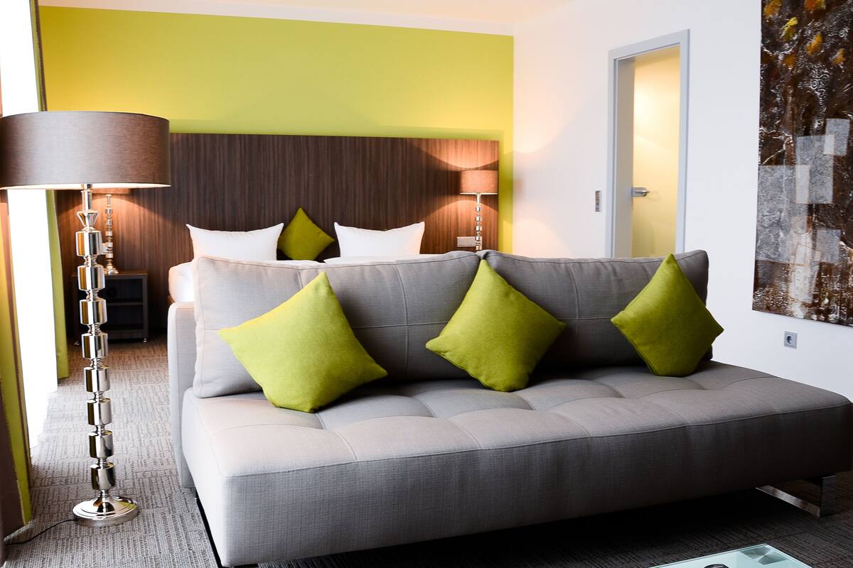 A modern hotel room with a grey sofa, green cushions, a standing floor lamp, and a large abstract painting on the wall.