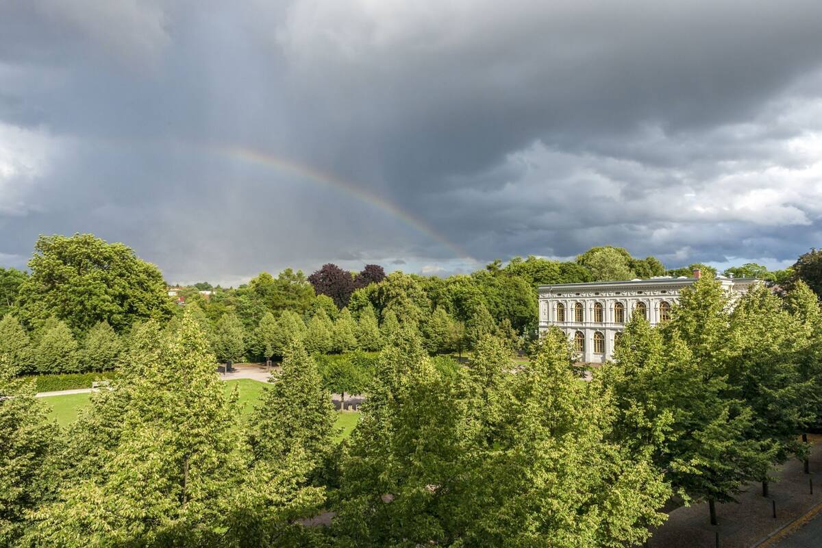 A large building with classical architecture surrounded by dense green trees and a visible rainbow.