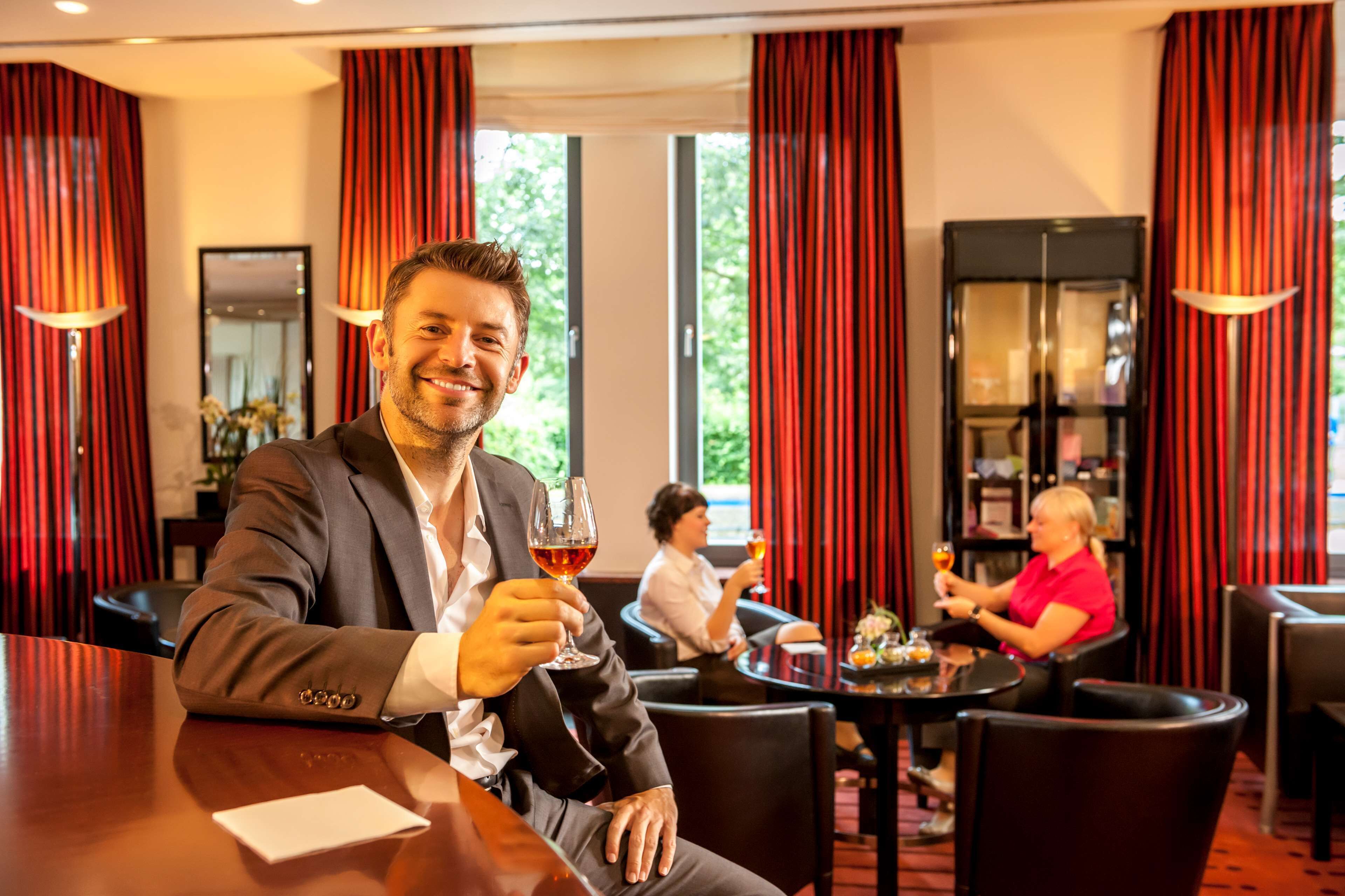A man in a suit holding a glass of wine at a table in a hotel lobby with red curtains and a large window.