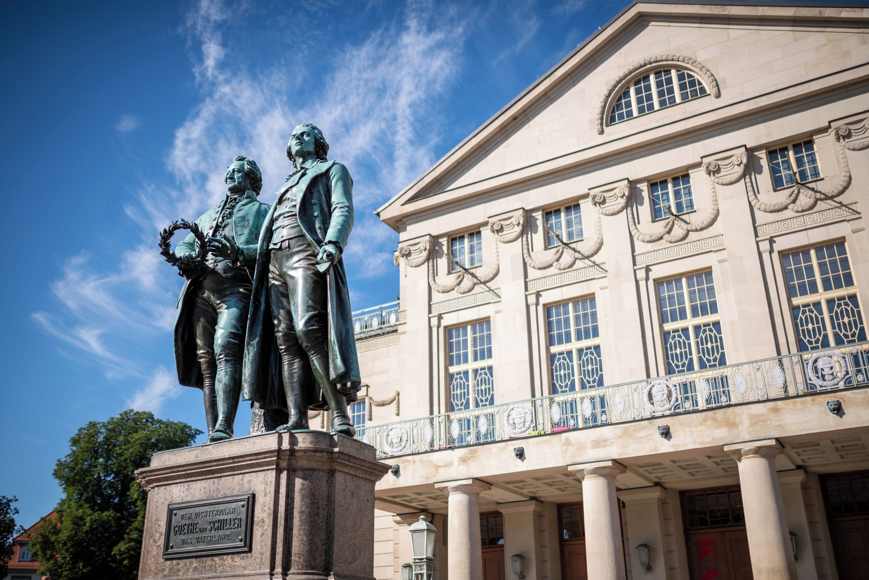 En staty som föreställer två figurer, placerad framför en klassisk byggnad med kolonner och dekorativa fönster.