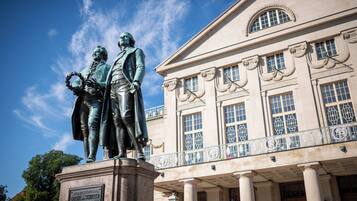 En staty som föreställer två figurer, placerad framför en klassisk byggnad med kolonner och dekorativa fönster.