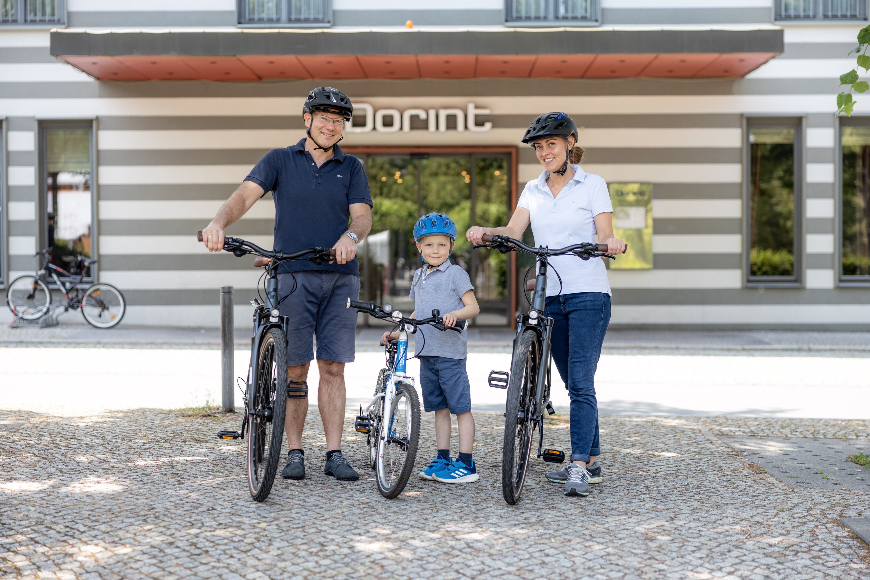 A family with bicycles in front of a building with a striped facade and the word "Dorint" visible.