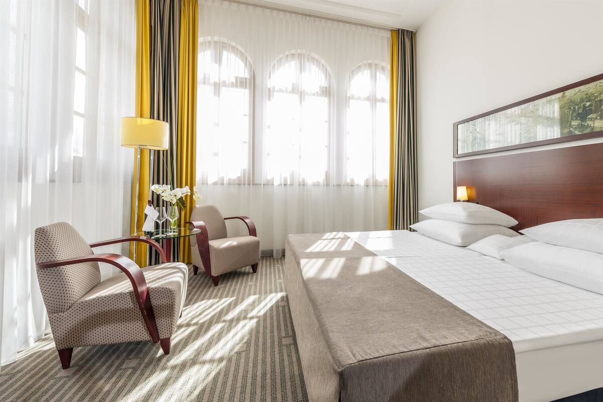 A hotel room with a bed, two armchairs, a small table with a lamp, and large windows with curtains.